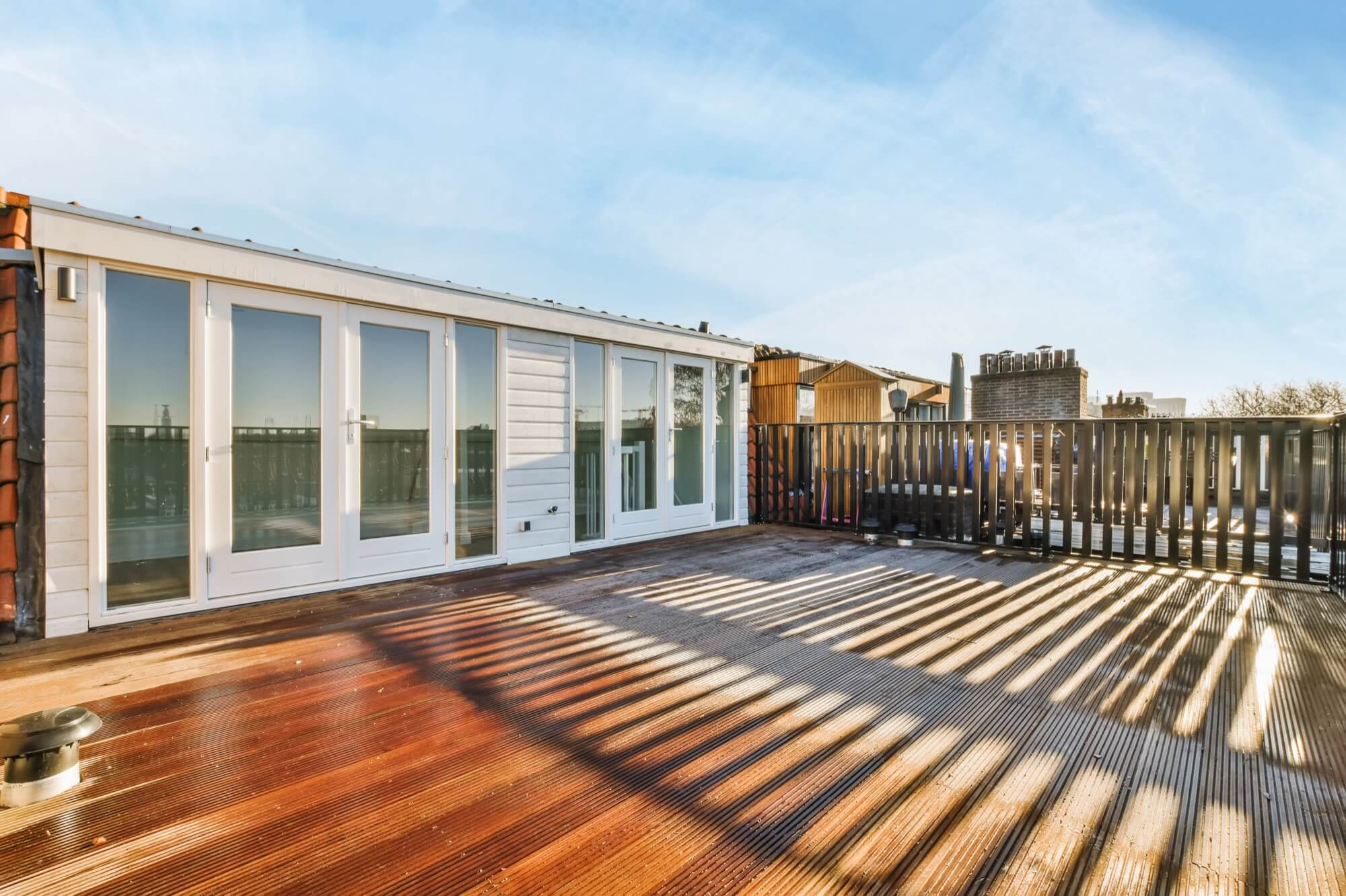 Sunny roof balcony with white double doors and railings that cast shadows over the wooden decking.
